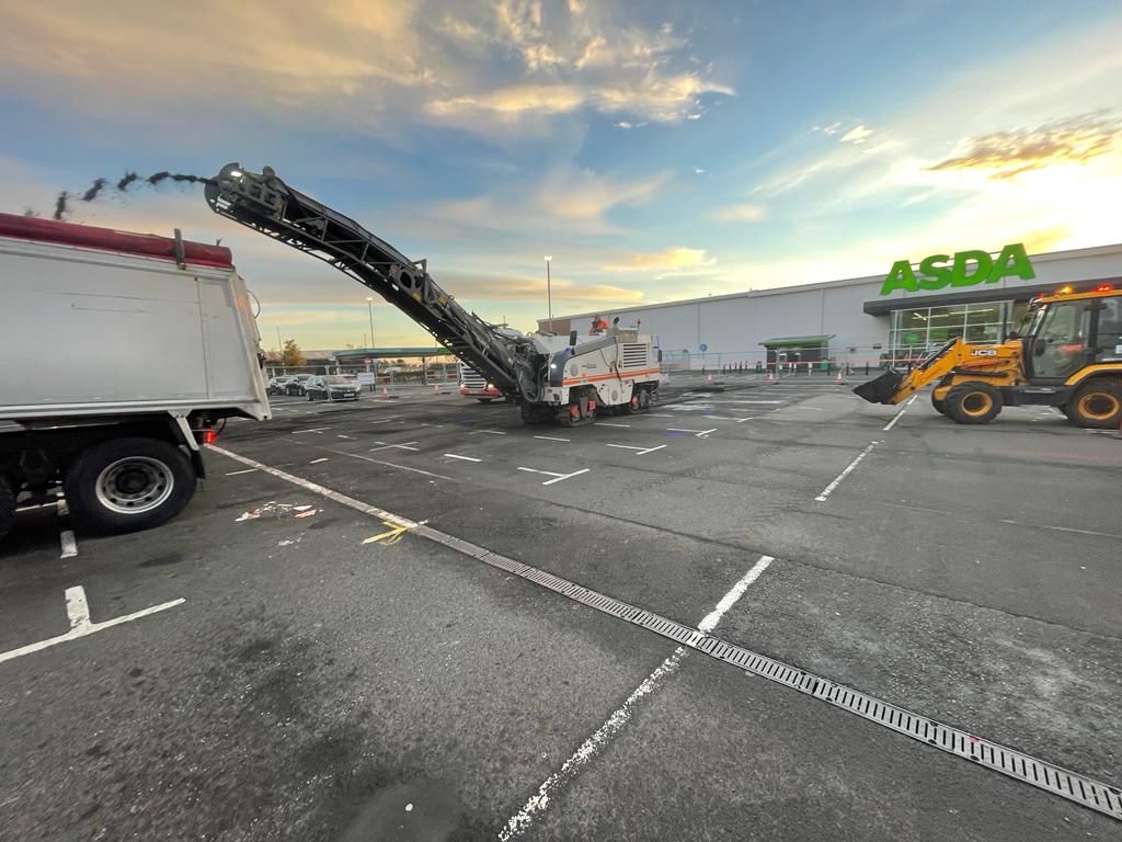 Cold planer and backhoe loader working across the car park at dusk with a tipper alongside.
