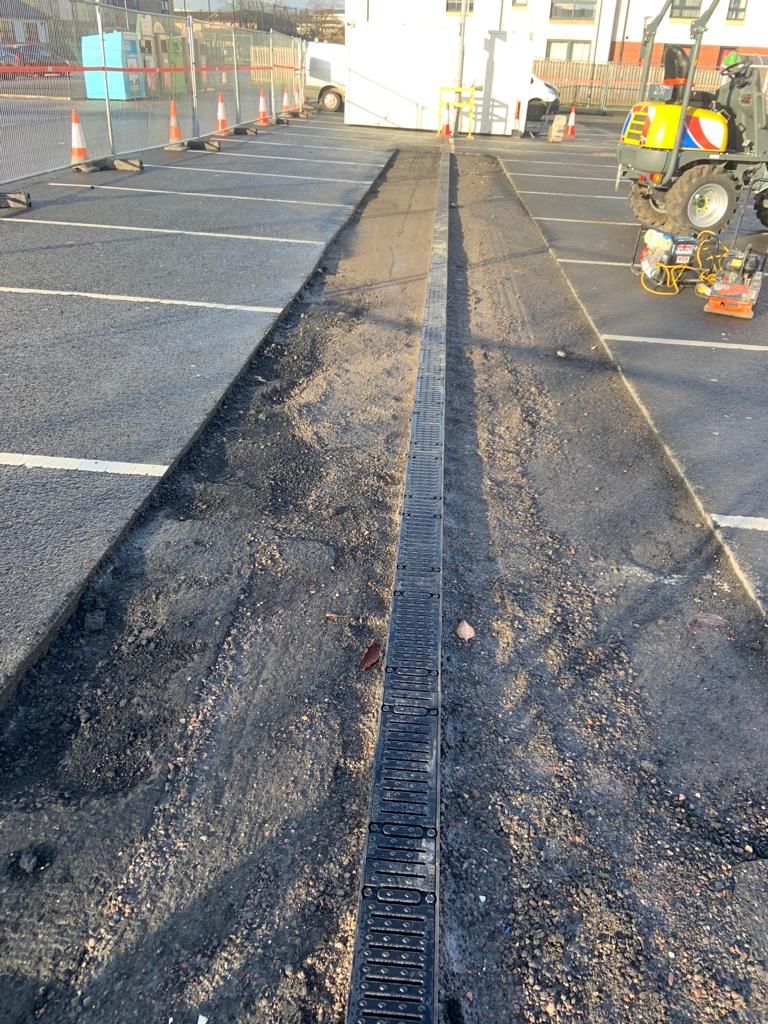 Long run of new drainage channel through freshly laid black asphalt, plant working at the far end.